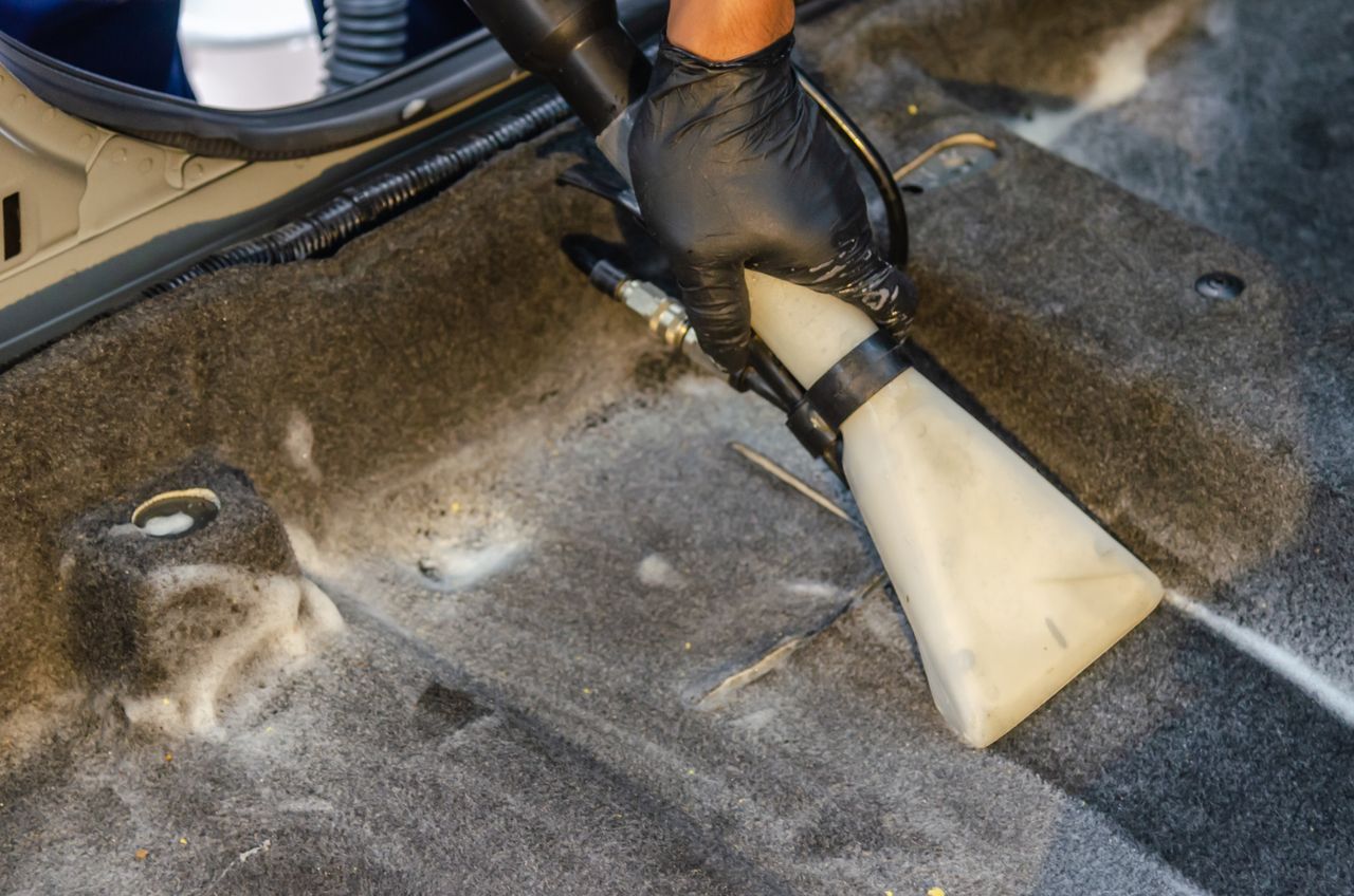 services-05 A hand in a glove using a vacuum to clean a car interior carpet, removing dirt effectively.