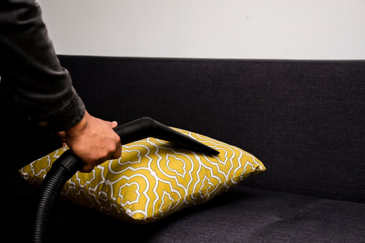 services-09 Close-up of a person vacuuming a patterned pillow on a dark sofa indoors.