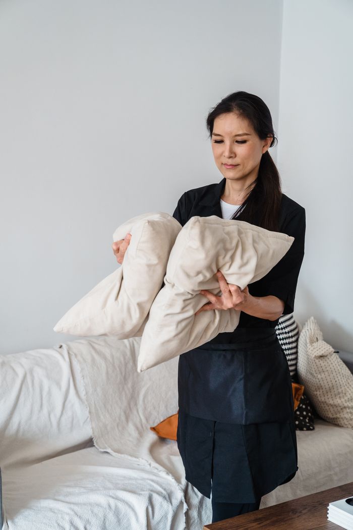 about-02 Smiling housekeeper arranging pillows on a sofa in a bright living room.